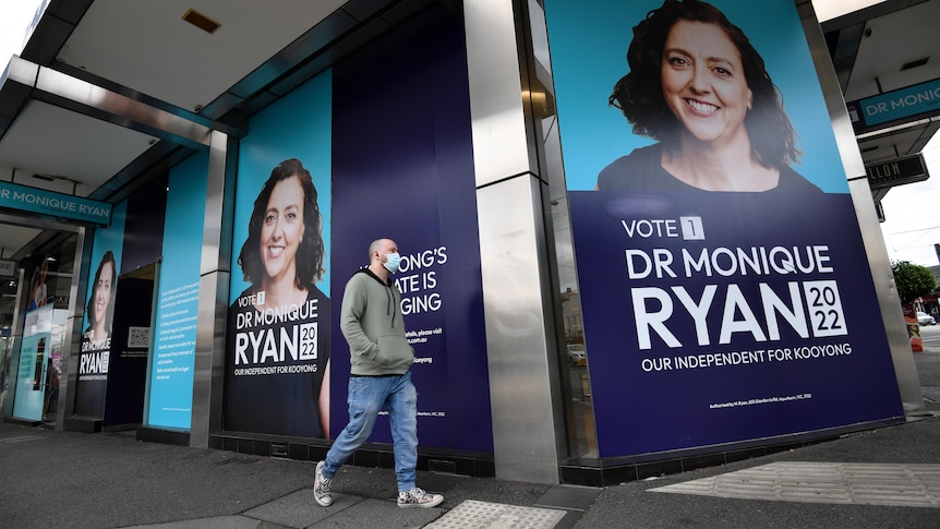 A man wearing a mask walks past large posters of Kooyong candidate Monique Ryan on a building.