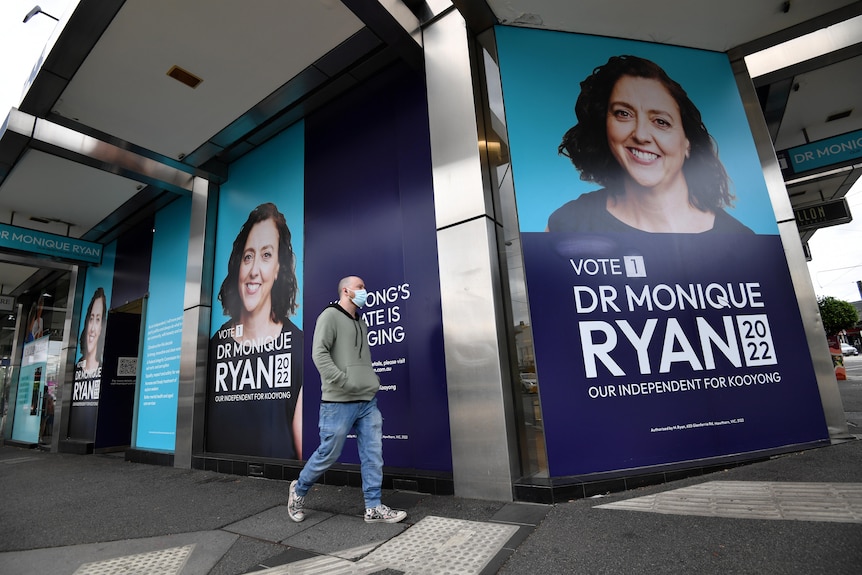 A man wearing a mask walks past large posters of Kooyong candidate Monique Ryan on a building.