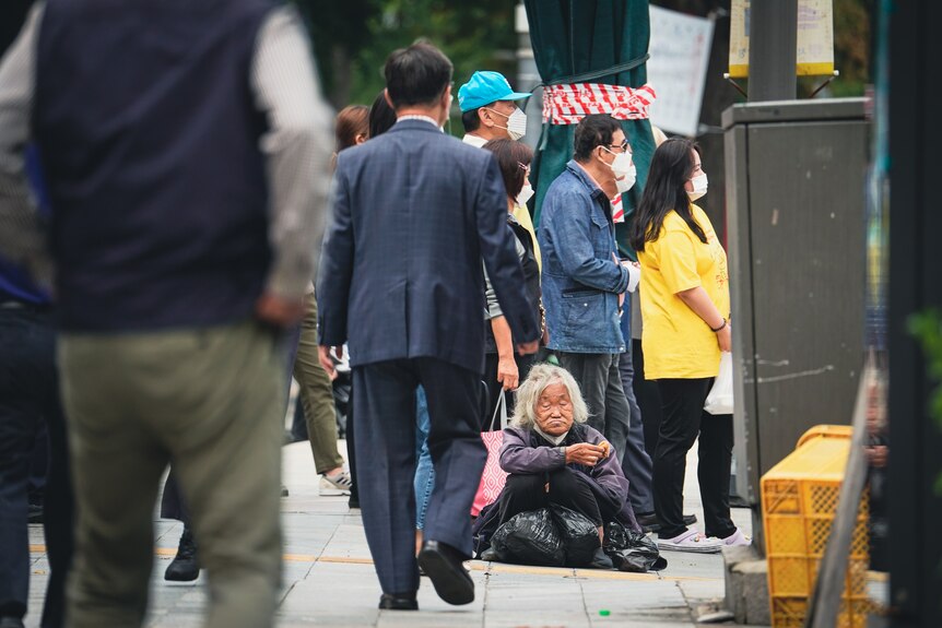 A man in a suit walks past an elderly woman sitting on a street corner