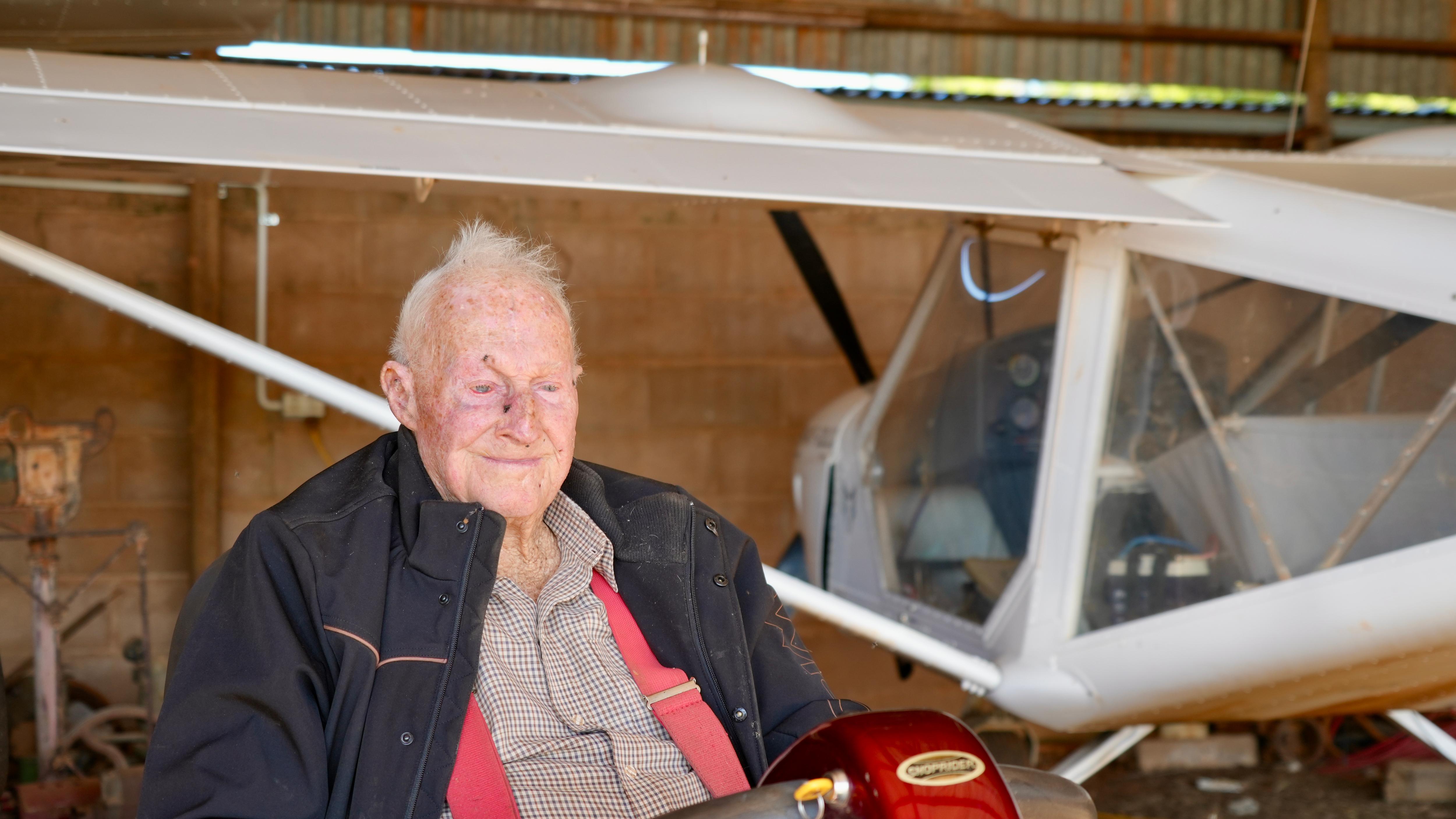 an old man with grey hair and a black jacket smiles next to a small white aircraft in a shed