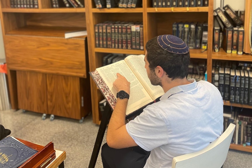Person seen from behind reading large book, with short hair and dark top, sits in library. He wears Jewish skullcap.