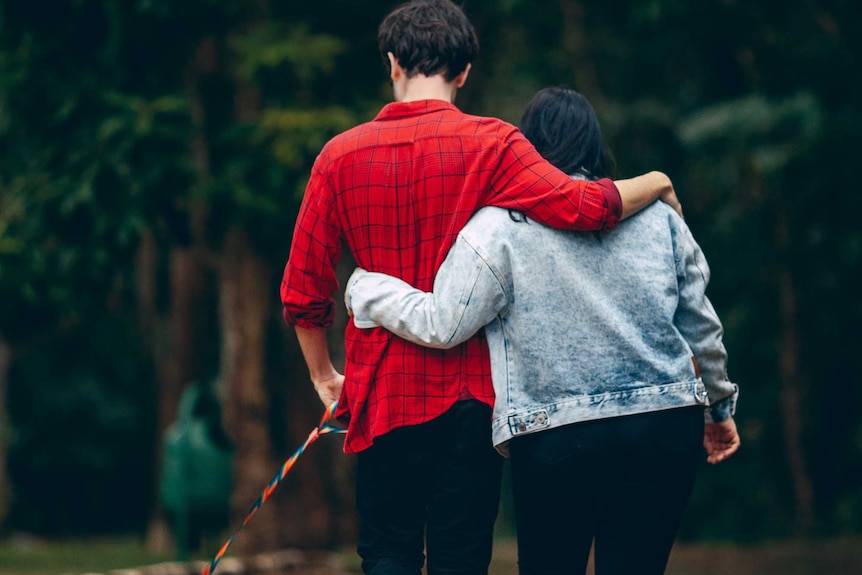 Young woman with hand wrapped around her boyfriend walks in a park with a dog.