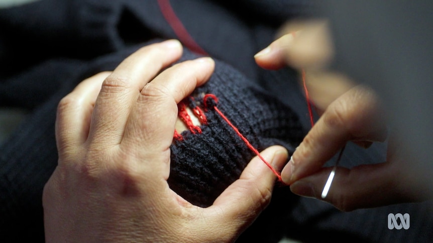 A hand holds a needle and thread to repair a hole in a jumper
