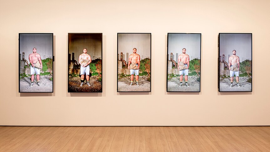 A topless man in denim shorts stands and holds a large rock at different times of day across 5 vertical video screens.
