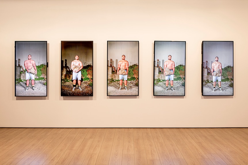 A topless man in denim shorts stands and holds a large rock at different times of day across 5 vertical video screens.