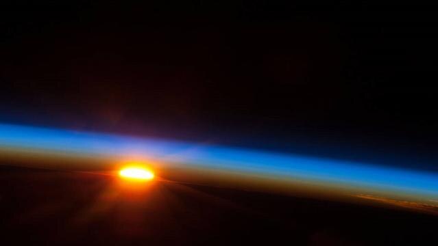 Sun peaks above Earth horizon with thin line of atmosphere above
