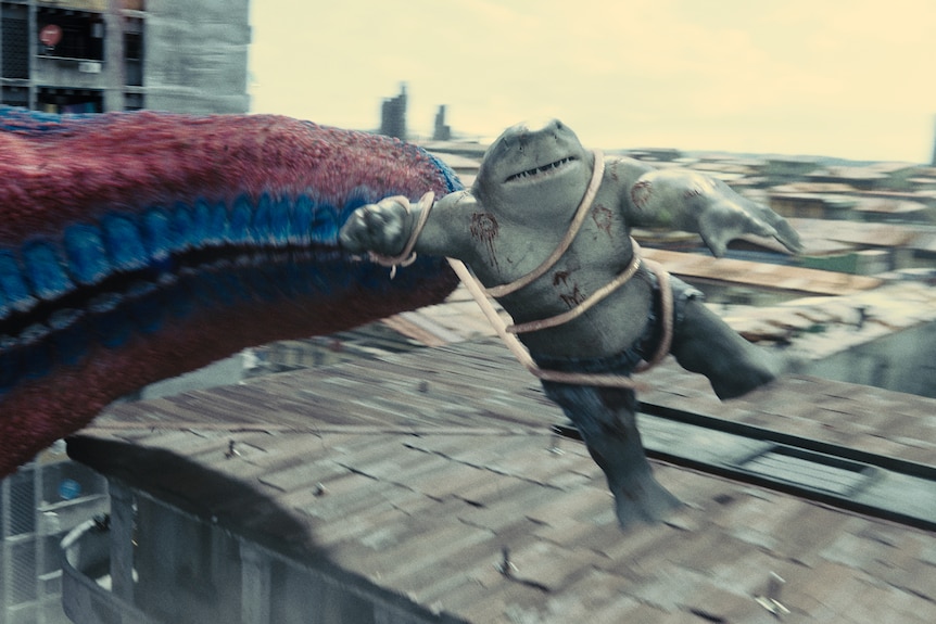 An anthropomorphised shark with arms and legs is pushed off the roof of a building by a giant monster arm