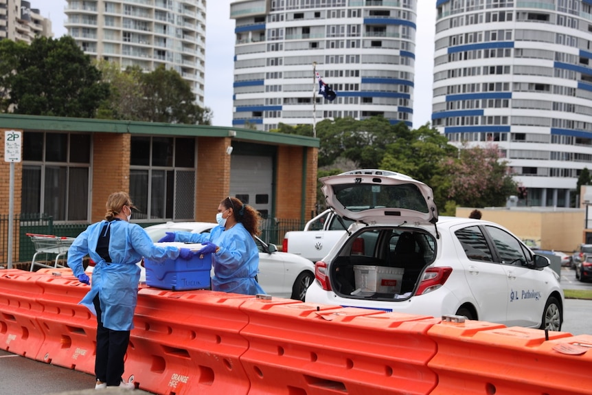 Medical staff dressed in blue coats, gloves and masks handing a blue esky over orange road barricades