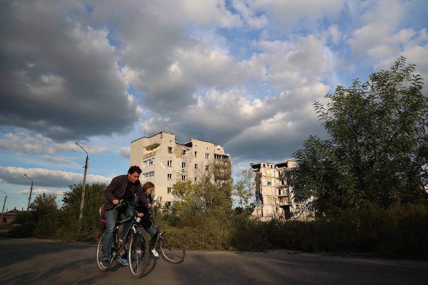 People ride bikes in front of a smashed apartment building.