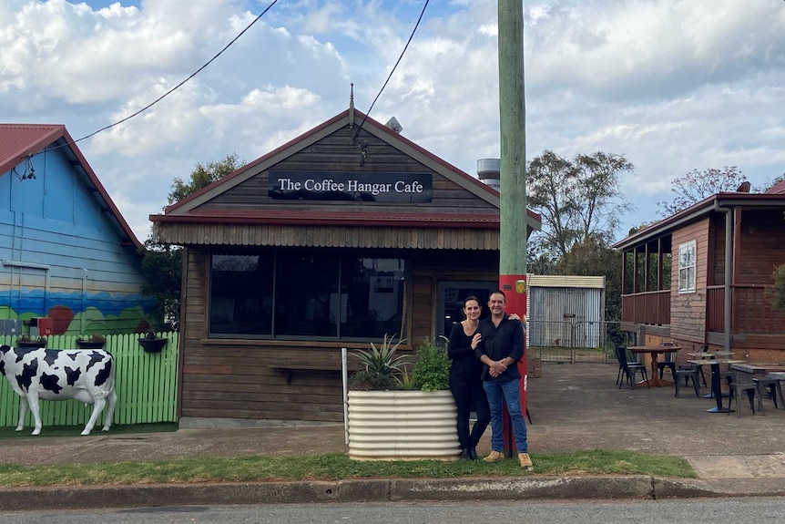 A man and woman stand together in front of a small wooden cafe with the sign, 'Coffee Hangar Cafe'