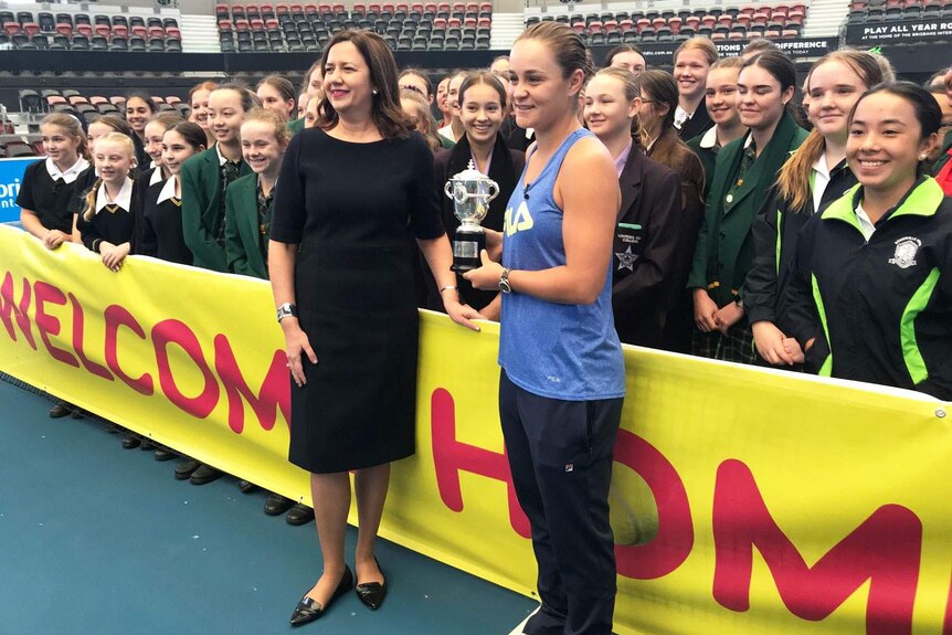 Tennis player Ash Barty holds a trophy as she stands with Premier Annastacia Palaszczuk with a crowd of students behind them.