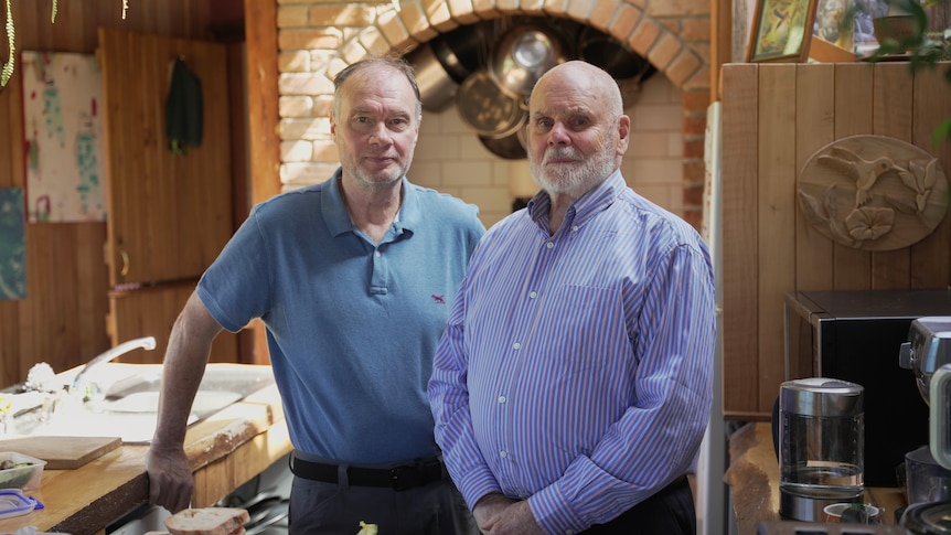 Two men stand in a kitchen with freshly prepared sandwiches on the bench in front of them