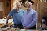 Two men stand in a kitchen with freshly prepared sandwiches on the bench in front of them