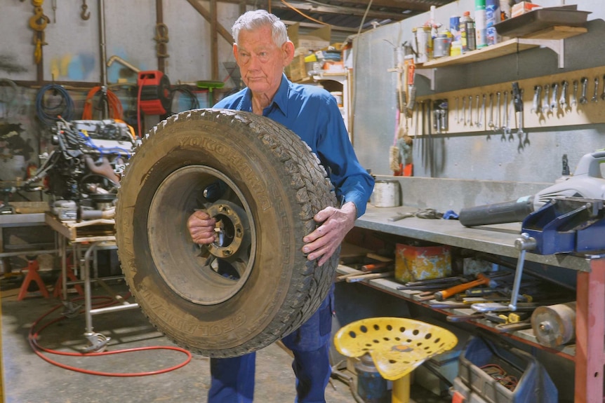 An elderly man carries a car tyre