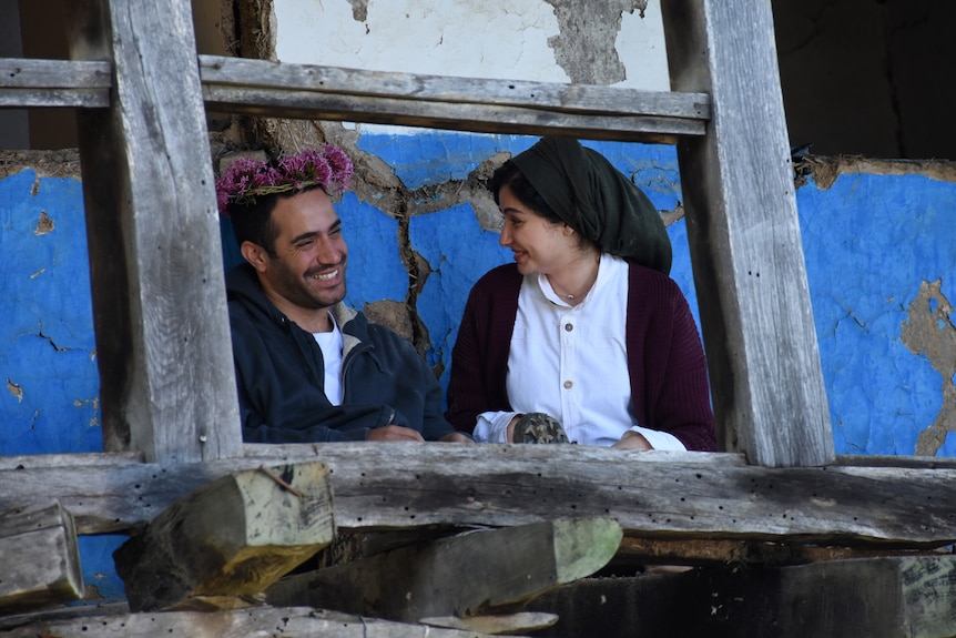 Jeune homme et femme s'asseoir ensemble en riant ;  il a une couronne de fleurs roses sur la tête, elle porte un foulard noir.