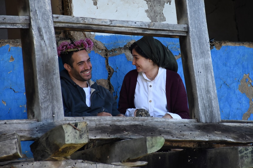 Young man and woman sit together laughing; he has a crown of pink flowers on his head, she is wearing a black head scarf.