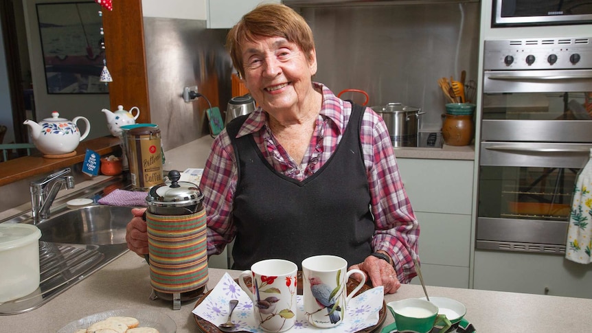A woman in a checked shirt and blue vest serves coffee and biscuits in a kitchen.