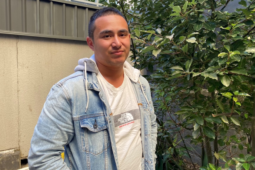 A man with short brunette hair, white tshirt and denim jacket stands smiling into the camera.