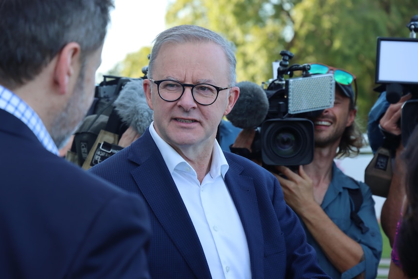 A man with dark glasses wearing a blue suit looks serious. He is surrounded by cameras.