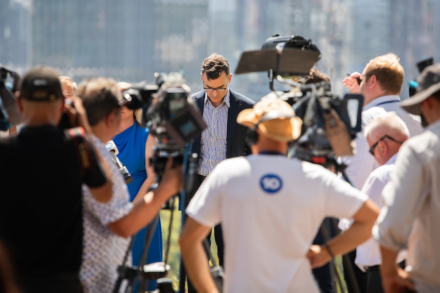 A man in a striped business shirt and blazer looks down at the ground, with a crowd of media in front of him.