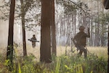 Two Ukrainian soldiers, one with a rocket launcher, walk through a sunlight clearing in the woods.