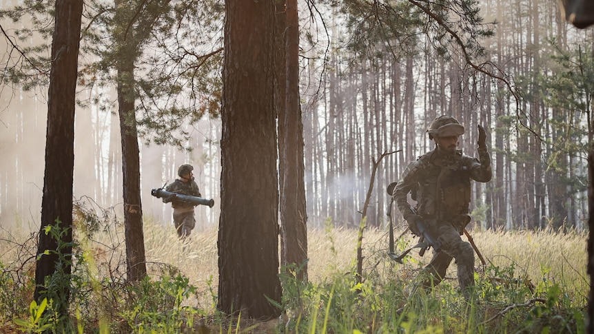 Two Ukrainian soldiers, one with a rocket launcher, walk through a sunlight clearing in the woods.