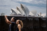 man and woman taking photo in front of a black fence with opera house in background