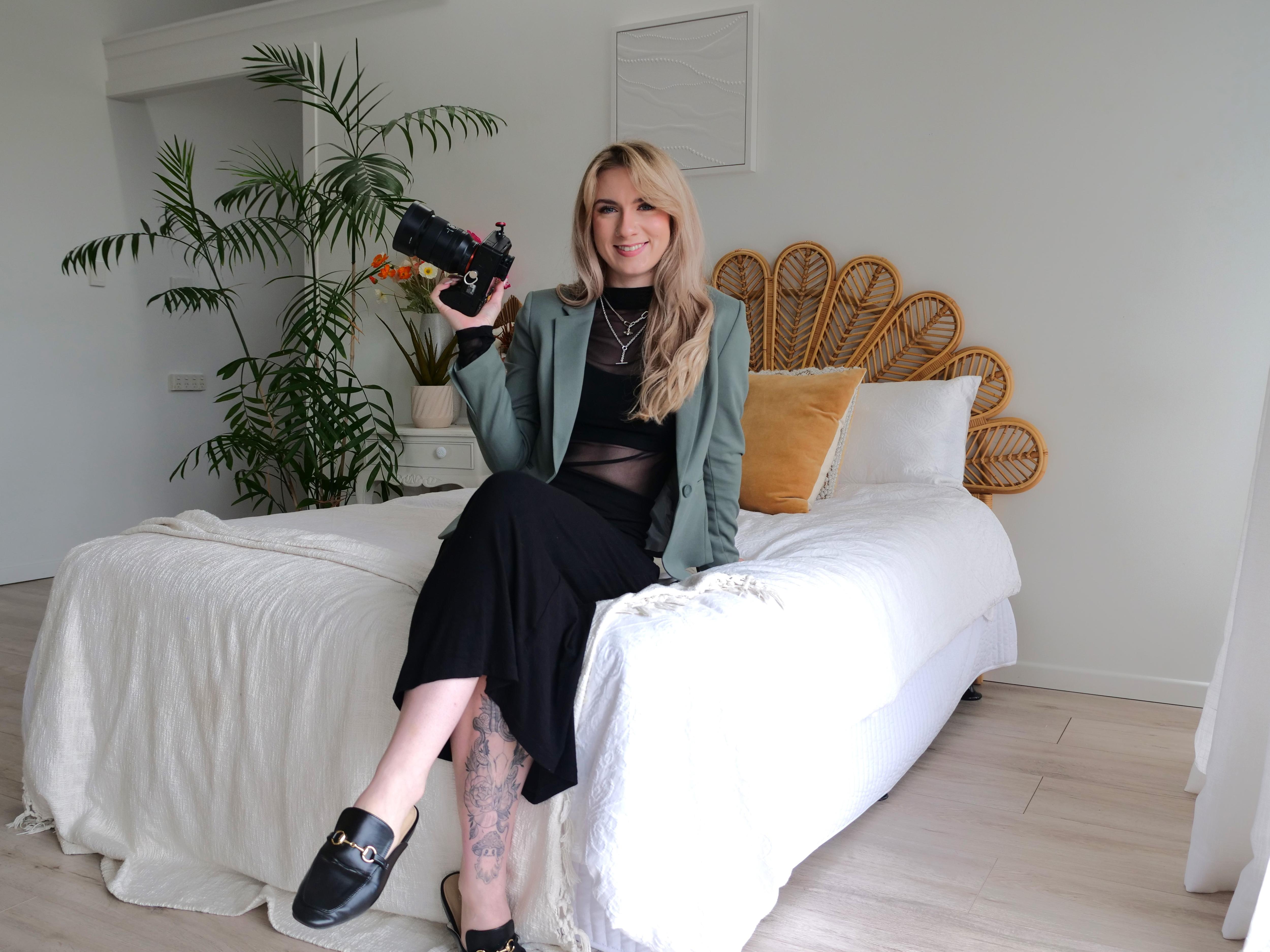 Young woman dressed in black holding a large camera, sitting on a bed and smiling. 