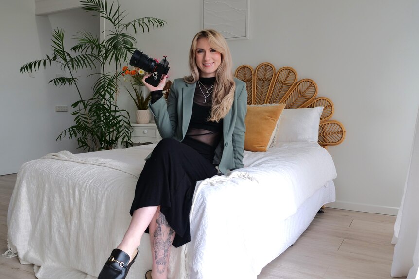 Young woman dressed in black holding a large camera, sitting on a bed and smiling. 