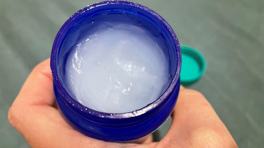 A hand holds an open jar of vapour rub, with the lid in the background. 