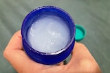 A hand holds an open jar of vapour rub, with the lid in the background. 