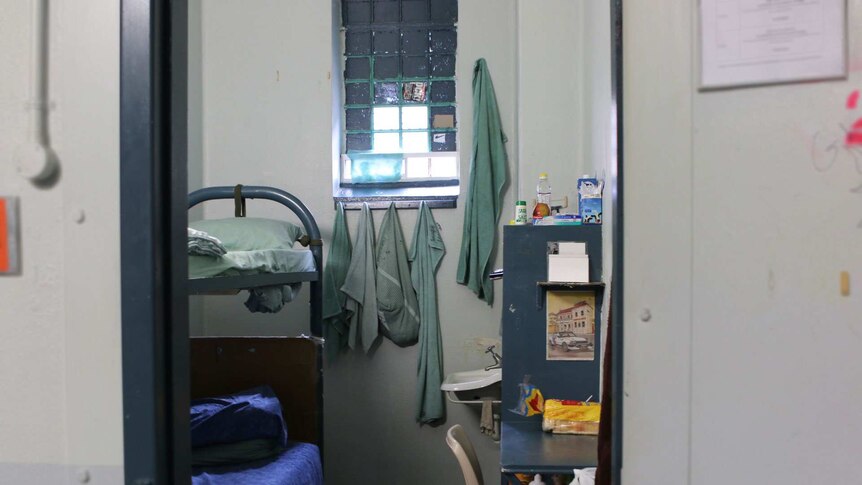 Through the door of a jail cell, a double bunk and a table and shelves, and a loaf of white bread can be seen.