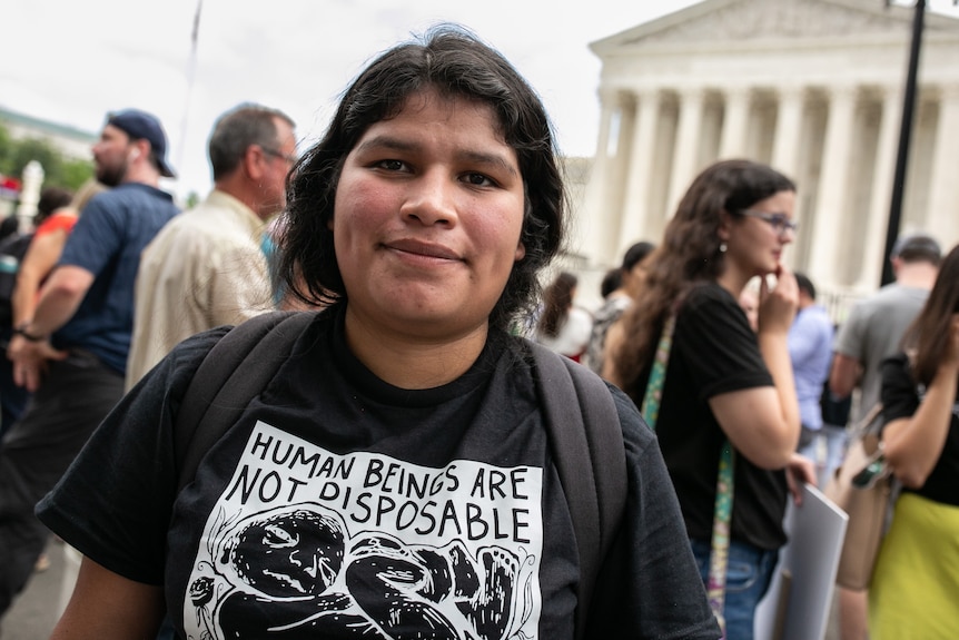 A brunette woman in a black t-shirt reading "human beings are not disposable" stands in front of the Supreme Court