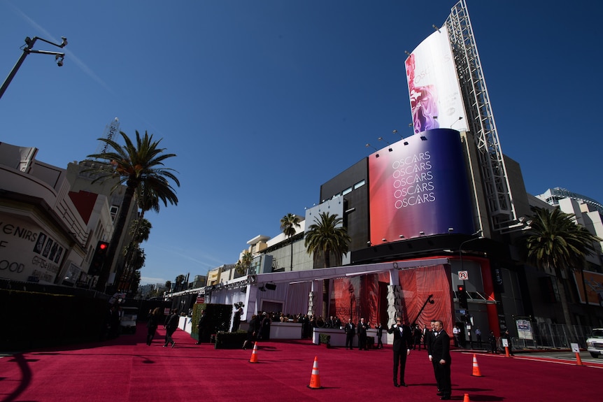 Security guards stand on red carpet outside a white-curtained area with people in formal wear.