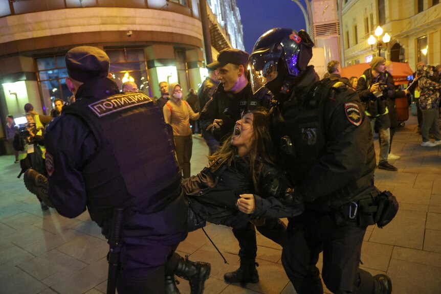 A woman yells as she is lifted and carried away by three police in tactical gear. 