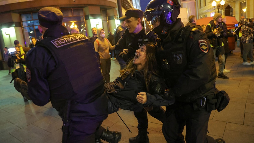 A woman yells as she is lifted and carried away by three police in tactical gear. 