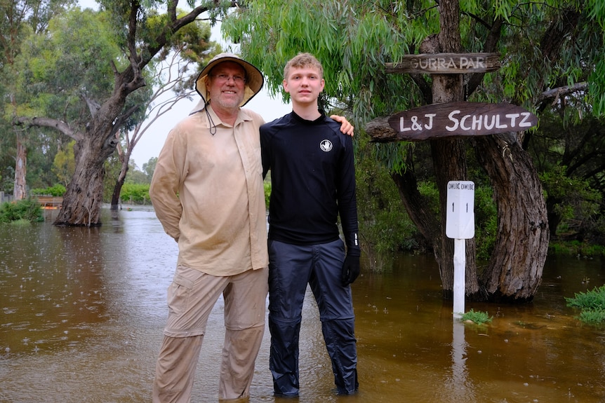 An older man and a younger man standing in flood water among trees and a sign saying L&J Schultz