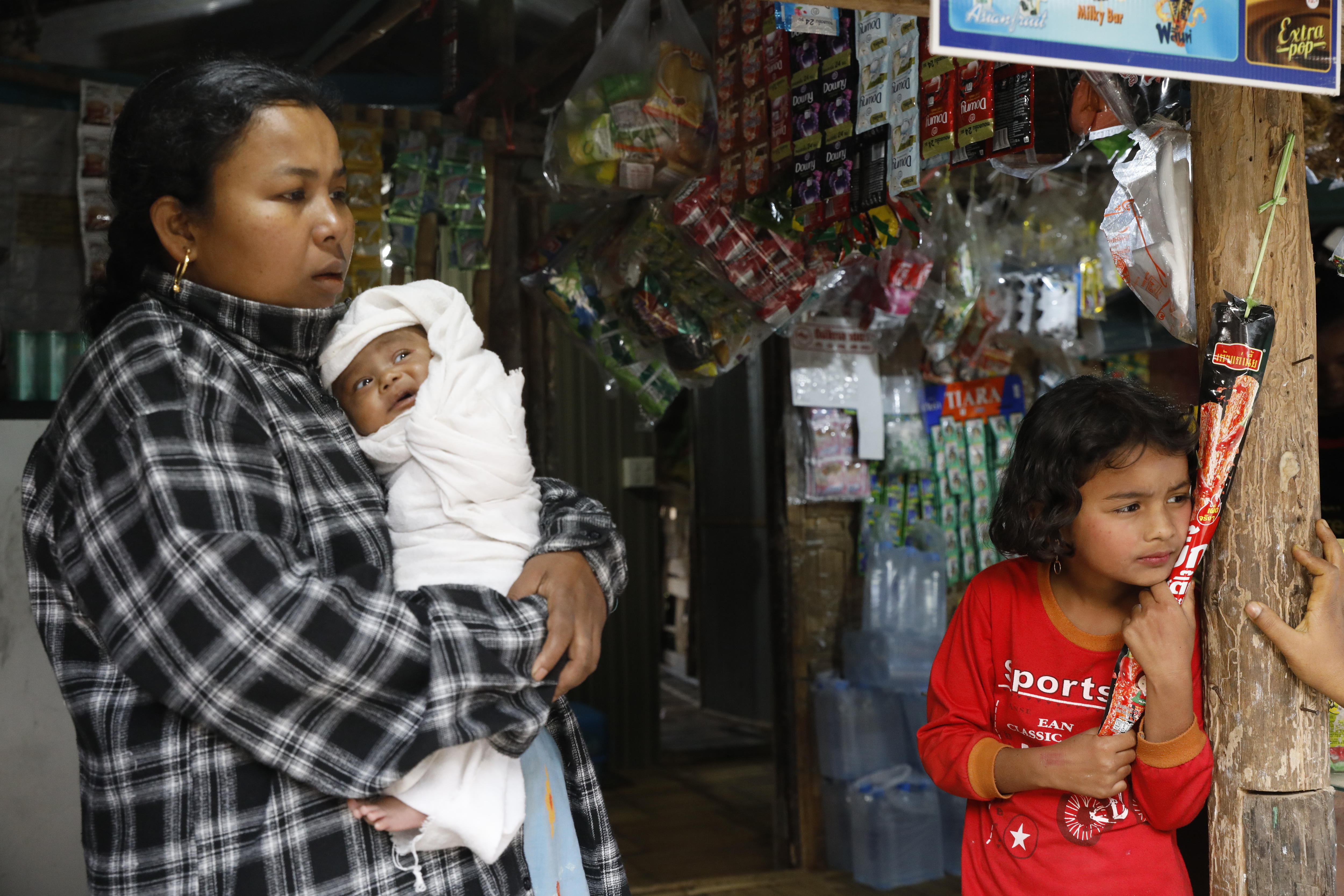 A mother holds a swaddled infant while her young daugher stands nearby leaning on a timber post.