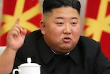 Kim Jong-un holds his right index finger up while speaking. He's at a desk with flags behind him.