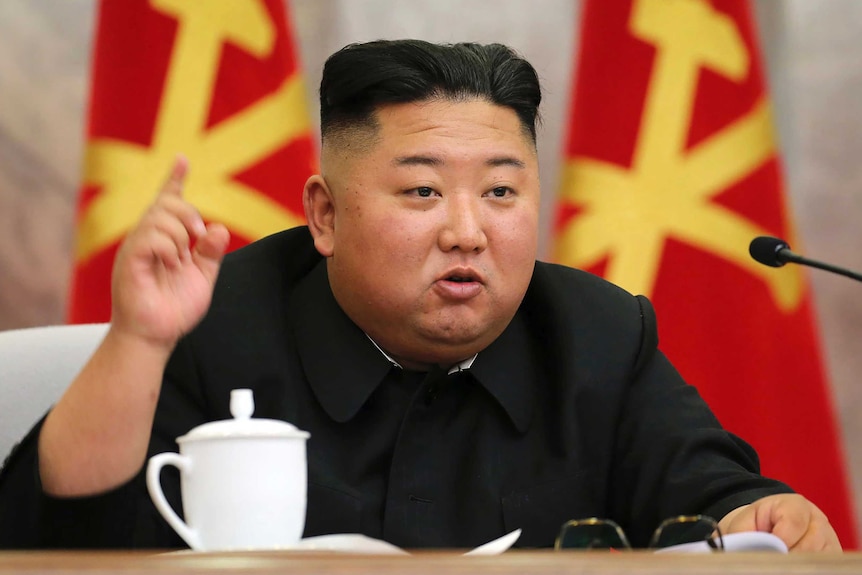 Kim Jong-un holds his right index finger up while speaking. He's at a desk with flags behind him.