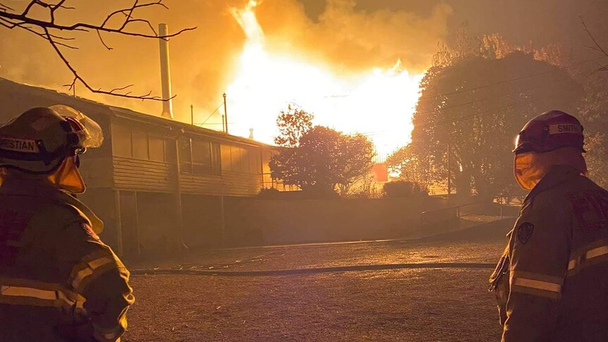 Two firefighters look on as a weatherboard building is alight in large flames tens of metres in height.