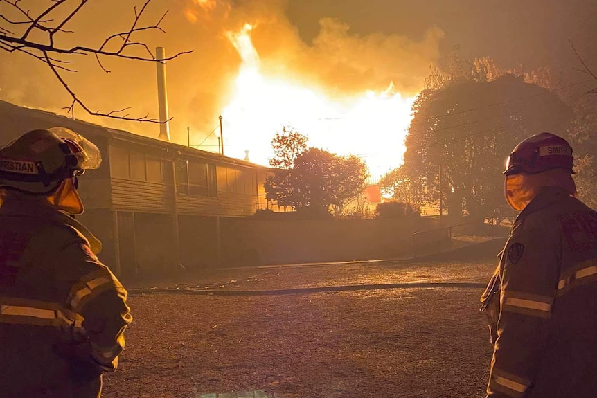 Two firefighters look on as a weatherboard building is alight in large flames tens of metres in height.