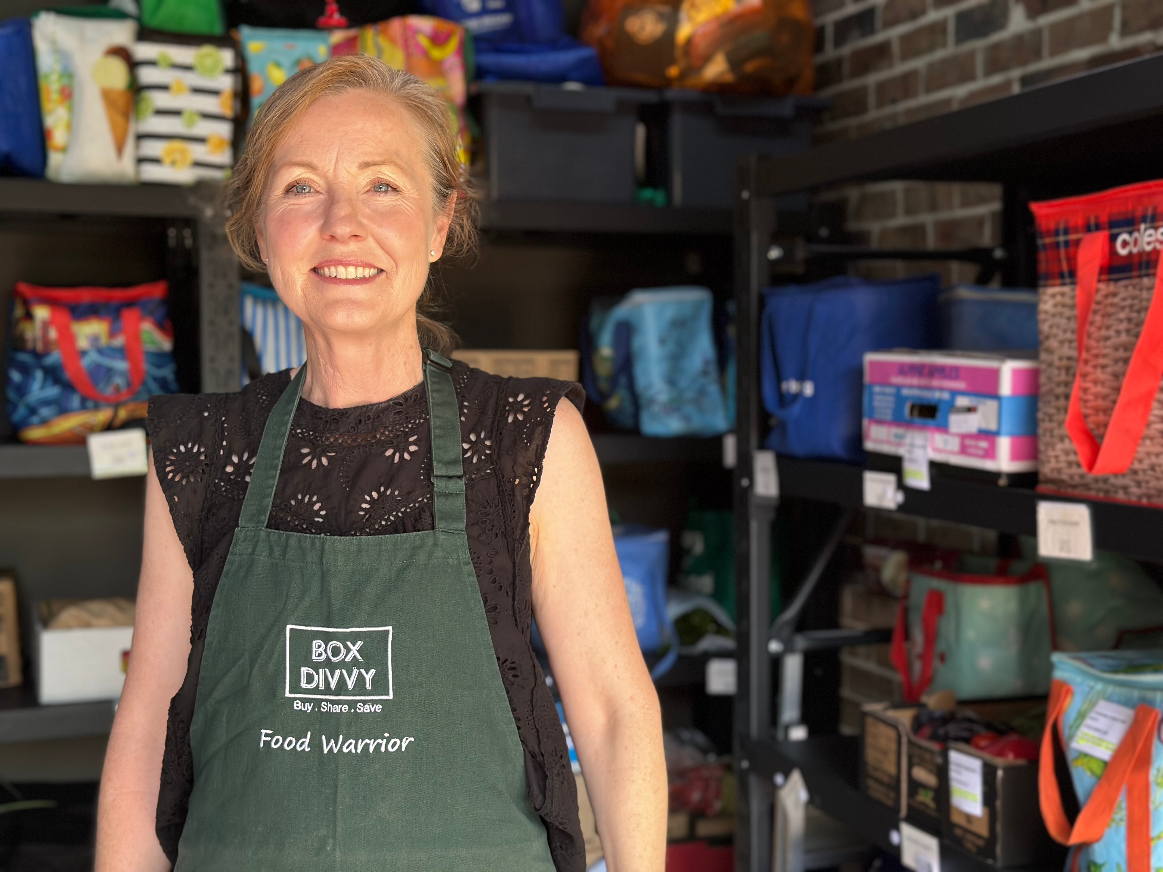 Una mujer mira a la cámara parada frente a un estante lleno de cajas con verduras y frutas. 