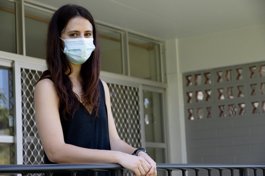 A woman wearing a surgical mask standing on the balcony of an apartment. 