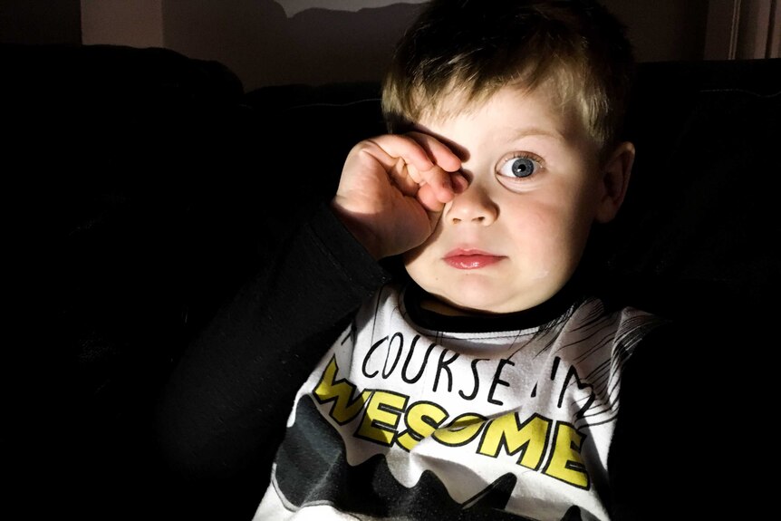 A young boy's face is lit up by an electronic device as he rubs his eye