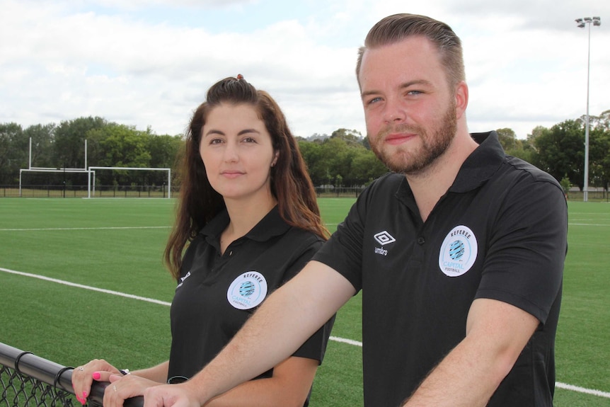 A female and male referee stand on the side of a soccer field.