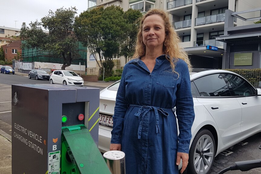 A woman stands in front of a car charging in an electric car station near apartments