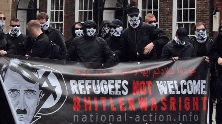 Masked National Action members hold a banner with an image of Hitler. It reads: 'Refugees not welcome Hitler was right'.