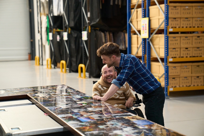 Daniel leans over to observe a table covered in photographs as David laughs next to him.