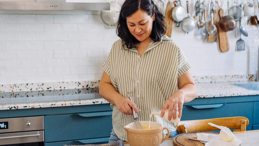 Emiko stands in a blue kitchen with white tiles and terrazzo counter looking down as she whisks something with a smile.